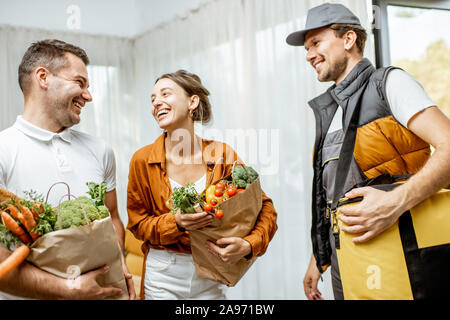 Fröhlich Kurier in Uniform liefert frische Lebensmittel zu einer jungen Familie zu Hause. Online Shopping und Home delivery Konzept Stockfoto