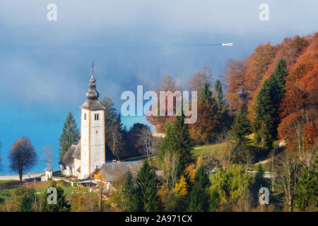 Herrliche Aussicht der katholischen Kirche von Johannes dem Täufer am Bohinjer See im Herbst und Kanu aus Nebel an einem sonnigen Morgen in Slowenien. Reisen, t Stockfoto