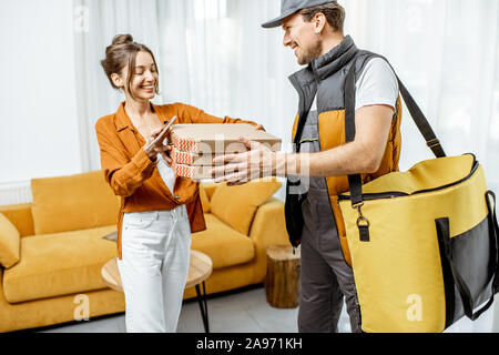 Stattliche Kurier in Uniform liefern Pizza mit thermischen Beutel an einen Client zu Hause, glücklich, Frau ihre Bestellung empfangen Stockfoto