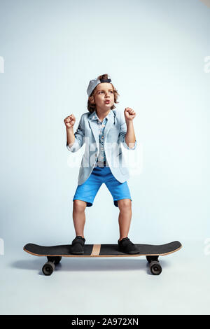Hübsche junge Junge auf Skateboard in Freizeitkleidung auf weißem Studiohintergrund. Reiten und sieht glücklich aus. Kaukasier Männlich Vorschüler mit hellen Gesichts Emotionen. Kindheit, Ausdruck, Spaß zu haben. Stockfoto