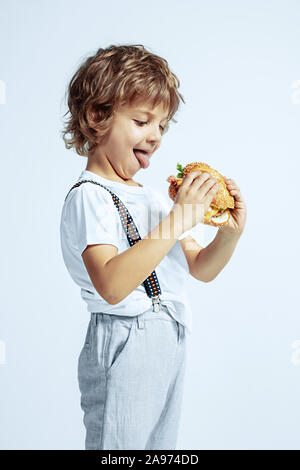 Hübsche junge lockige Junge in Freizeitkleidung auf weißem Studiohintergrund. Essen Burger. Kaukasier Männlich Vorschüler mit hellen Gesichts Emotionen. Kindheit, Ausdruck, Spaß, fast food. Stockfoto