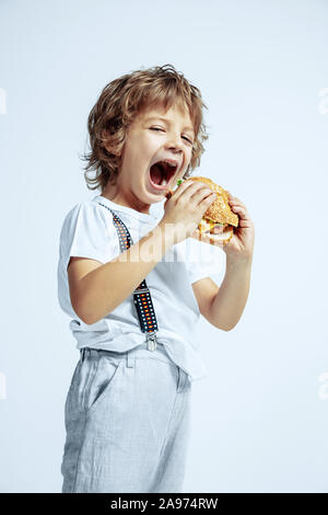 Hübsche junge lockige Junge in Freizeitkleidung auf weißem Studiohintergrund. Essen Burger. Kaukasier Männlich Vorschüler mit hellen Gesichts Emotionen. Kindheit, Ausdruck, Spaß, fast food. Stockfoto