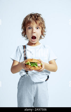 Hübsche junge lockige Junge in Freizeitkleidung auf weißem Studiohintergrund. Essen Burger. Kaukasier Männlich Vorschüler mit hellen Gesichts Emotionen. Kindheit, Ausdruck, Spaß, fast food. Erstaunt. Stockfoto