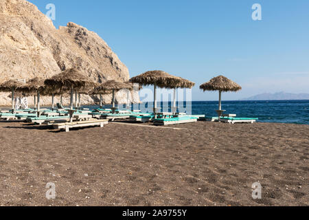 Liegestühle mit Stroh Sonnenschirme auf den Black Sand Beach in Perissa in Santorin, Griechenland. Stockfoto