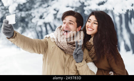 Glückliches Paar unter Selfie am Telefon lächeln und gestikulierend Sieg Zeichen stehen im verschneiten Winterwald. Panorama Stockfoto