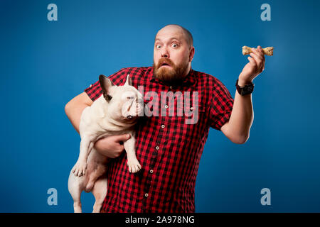 Überrascht Mann im Hemd hält Bulldogge und Knochen auf leeren blauen Hintergrund im Studio Stockfoto