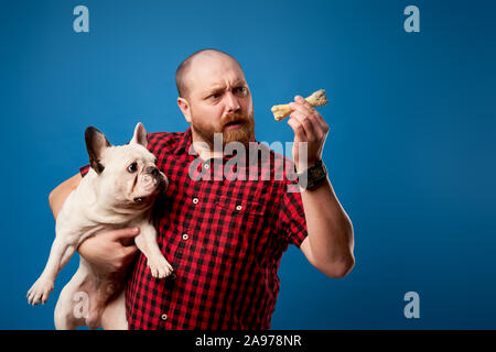 Mann im Hemd hält Bulldogge und Knochen auf leeren blauen Hintergrund im Studio Stockfoto