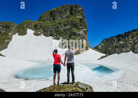 Ein Paar genießt die Aussicht einer gefrorenen Bergsee. Das Eis scheint in vielen verschiedenen Blautönen. See ist von hohen Bergen umgeben. Kein Schnee auf der Stockfoto