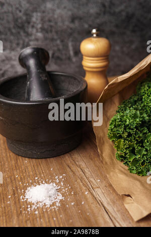 Anordnung von Kale in Brown Paper Bag, schwarz Mörser und Stößel, Holz- Pfeffer Mühle, Haufen von grobem Meersalz auf Holz- Oberfläche, dunklen Hintergrund Stockfoto