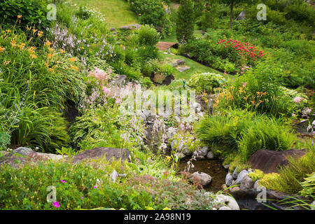 Blick von oben auf die schräge Wohn- Garten im Hinterhof mit Felsen umrandete Stream umgeben von orange Hemerocallis - Daylily, rosa Anemone hupehensis Blumen, etc. Stockfoto