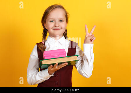 Closeup Portrait von einem kleinen Mädchen Schülerin auf einem gelben Hintergrund. Das Kind hat ein Lunchpaket und ein Buch in der Hand, sie zeigt ein Sieg unterzeichnen. Zurück Stockfoto