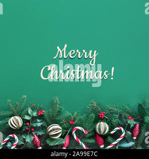 Kreative Weihnachten flach auf grünem Papier mit roten Verzierungen. Quadratische Komposition mit Begrüßungstext "Frohe Weihnachten!". Natürliche Tanne und Holly Zweige Stockfoto