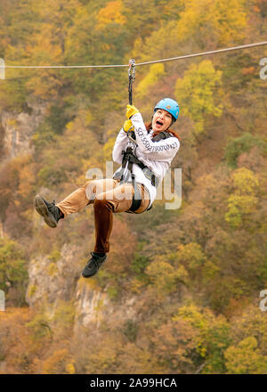 Eine Frau in einem Helm fliegt auf einer Seilrutsche über einen bunten Herbst Schlucht. Stockfoto