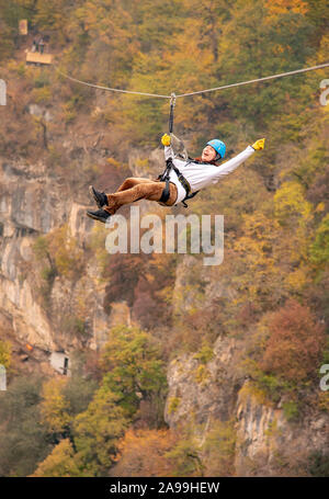 Eine Frau in einem Helm fliegt auf einer Seilrutsche über einen bunten Herbst Schlucht. Stockfoto