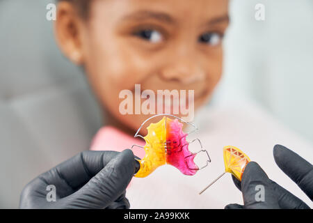 In der Nähe der Hände in schwarzem Gummi medizinische Handschuhe halten bunte Klammern. Zahnarzt mit Zahnspangen aus Metall für afrikanische lächelnde Mädchen. Konzept Korrektur und Verbesserung beißen, Gesundheit und Schönheit. Stockfoto