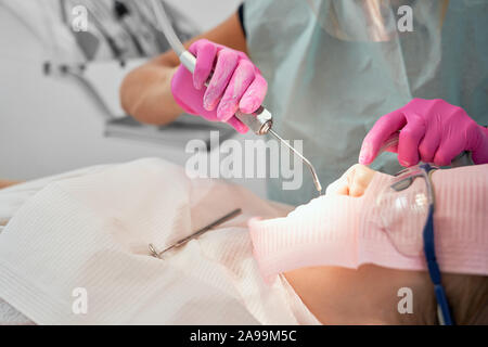 Patienten der zahnmedizinischen Klinik in zahnmedizinischen Stuhl mit weißem Latz und Schutzbrille. Zahnarzt in Gummihandschuhe Werkzeug zum Halten, gerade Zähne. Arzt tun Verfahren mit Dental curing UV-Licht. Stockfoto