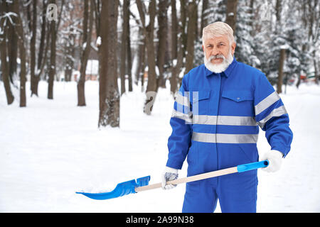 Ältere cleaner in Blau mit Sicherheit band Holding blau Schneeschaufel, posierend, an der Kamera schaut. Bärtige Arbeiter Reinigung Schnee auf der Park im Winter. Konzept der Stadt. Stockfoto