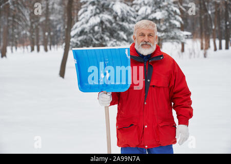 Porträt eines alten, bärtigen Reiniger mit blauer Schnee Schaufel auf dem Hintergrund der Natur. Hausmeister gekleidet in Winter Kleidung stehen im Freien, an der Kamera schaut. Stockfoto