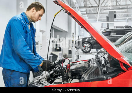 Seitenansicht der männlichen Ingenieur mit Licht Stoppeln, trägt blaue staubiger Arbeitskleidung und schwarzen Handschuhen, vor der geöffneten Motorhaube von rotes Auto in der Garage und mit Batterie Überbrückungskabel für Ladegeräte Stockfoto