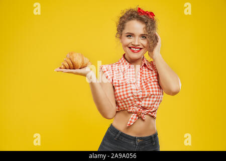 Lächelnd hübsche Frau mit roten Lippen und Lockige blonde Haare in Jeans Shorts gekleidet und kariertem Hemd Holding frische leckere Croissant auf ihre Hand und Kamera. Konzept der leckeren Snack und Vergnügen Stockfoto