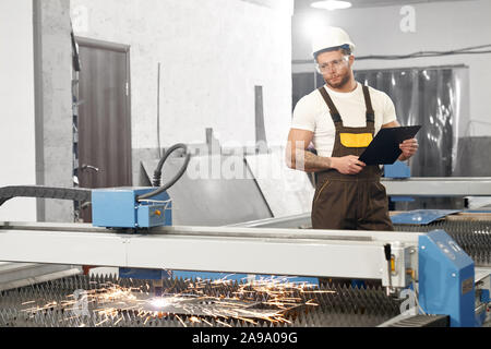 Ansicht von der Seite der ernster Mann in Schutzbrille und Helm, Ordner und Daten für die Steuerung von Geräten und Anlagen. Konzept der Plasma- und Metallarbeiten. Stockfoto