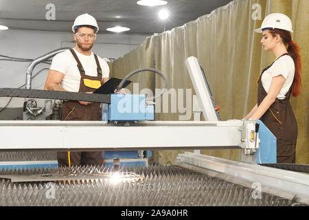 Ansicht von der Seite der professionellen weiblichen und männlichen Mechanik controlling Prozess der Plasmaschneiden auf Metall Werk. Team in Uniform und Helme Arbeiten mit Computern und CNC. Konzept der Zusammenarbeit. Stockfoto