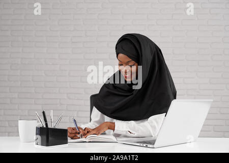 Vorderansicht des schönen weiblichen mit schwarzen Hijab lächelnd und schriftlich beim Sitzen am Tisch im Büro. Junge afrikanische Frau lächelnd und Spaß an der Arbeit im Unternehmen. Konzept der Business. Stockfoto