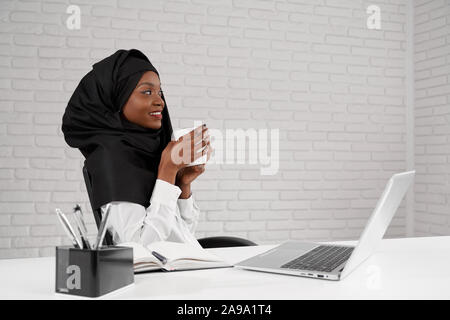 Seitenansicht der lächelnde Frau mit schwarzen hijab Kaffee trinken und Fenster, während Sie Pause am Job. Schöne afrikanische Dame in multi-ethnische Gesellschaft arbeiten. Konzept Kultur und Erfolg. Stockfoto
