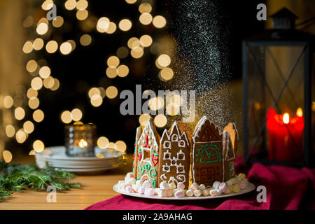 Kleine Lebkuchenhäuser mit Glasur auf Tisch mit roten Tischdecke und Dekorationen, Kerzen und Laternen. Lichter des Weihnachtsbaums Stockfoto
