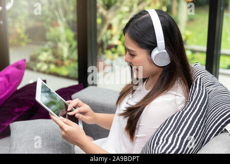 Ziemlich lächelnden jungen Frau tragen Kopfhörer sitzen auf dem Sofa und die Verwendung auf dem Tablet. Stockfoto