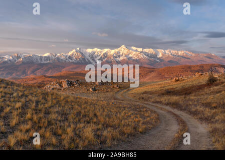 Schönen Herbst Sonnenaufgang mit einem gewundenen Feldweg auf einem Berg vor dem Hintergrund der Berge bedeckt mit Wald und Schnee, blauer Himmel und Cl Stockfoto