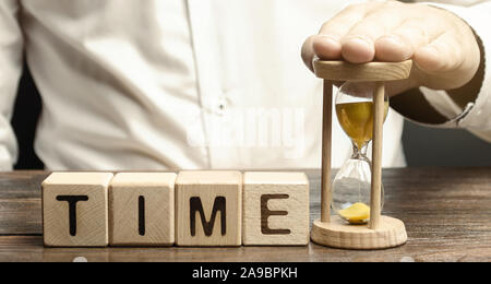 Ein Mann legt Sanduhr in der Nähe von Holz- Bausteine mit dem Wort Zeit. Konzept der Zeit Verwaltung und ordnungsgemäße Verteilung. Planung Wirtschaft und Arbeit. Business y Stockfoto