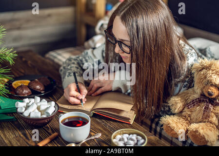 A young woman in a sweater is writing a wish list in a notebook with a mug of coffee in the evening in the warm atmosphere of Christmas. Cozy New Year Stockfoto