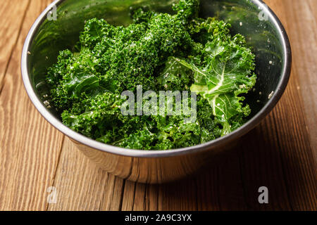 Zutaten für Kale Chips: Grünkohl mit Olivenöl, Salz, Sesam und Zitronensaft in Metall Schüssel gemischten auf strukturierte Holzoberfläche - Schrägansicht Stockfoto