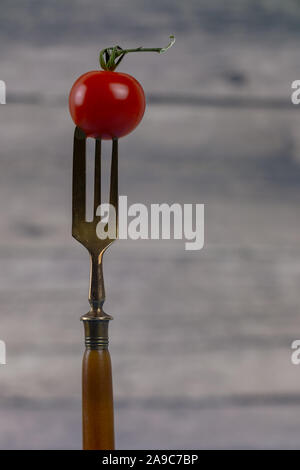 Kleine rote Tomate aufgespiesst auf einem antiken Gabel mit drei Zinken der Gabel auf unscharfen Holz- Hintergrund isoliert Stockfoto