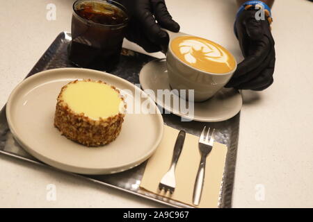 Barista mit schwarzen Handschuhen serviert Tablett mit Tasse Kaffee mit Blatt, Kuchen, Gabeln eingerichtet und schwarzen Kaffee in einem Glas Stockfoto