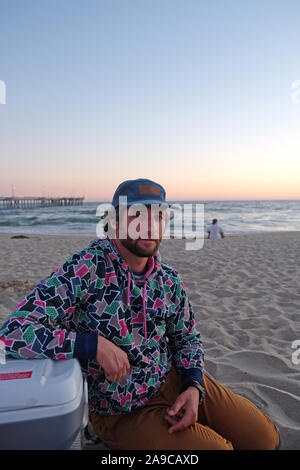 Mann mit Mütze mit Bart und lockiges Haar sitzt am Strand bei Sonnenuntergang Stockfoto