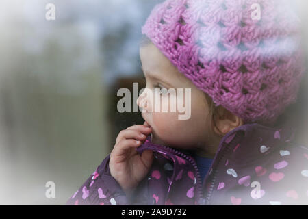 Mädchen mit rosa Mütze und Jacke stehen am Fenster suchen, den Finger auf den Mund, Kopf hoch Stockfoto