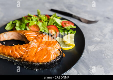 Gebratener Lachs Fisch Steak mit Gemüse und Salat Zitrone auf schwarze Platte. Kreative Gestaltung aus Fisch, Salat, Tomate, Gurke. Gesunde Ernährung Konzept Stockfoto