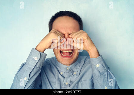 Ein Mann weint wie ein Kind. Ein junger Kerl in ein Hemd mit einem negativen Gefühl auf einem blauen Hintergrund. Stockfoto