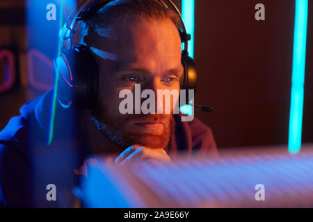 Junger bärtiger Mann Kopfhörer tragen und Computer Monitor mit nachdenklichen Blick er in dunklen Büro arbeiten Stockfoto