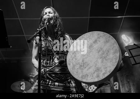 Eine weibliche Musiker ist von einem niedrigen Winkel gesehen wie sie singt und spielt Native American traditionelle Trommel, unscharfe band Mitgliedern gesehen werden, in den Hintergrund Stockfoto