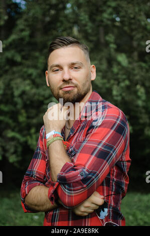 Brutale bärtiger junger Mann im roten T-Shirt steht im Freien auf grüne Bäume Hintergrund. Hüfthose style Konzept. Stockfoto