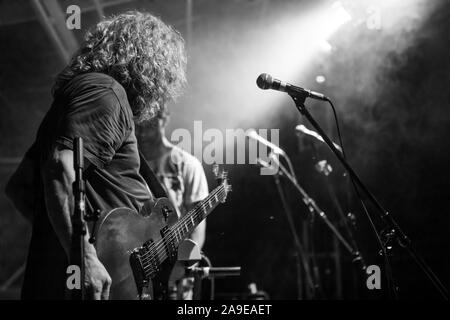 Ein Mann mit langen Haaren ist von Low Angle View angesehen, als er halten und spielen E-Gitarre, Schwarz und Weiß, mit den Bandmitgliedern im Hintergrund Stockfoto