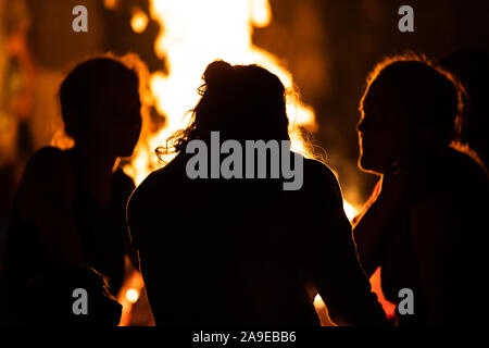 Silhouette der Gruppe von Personen, die vor dem Feuer sitzen wie sie reden, von hinten während der dunklen Nacht Lagerfeuer gesehen, verschwommenen Hintergrund Stockfoto