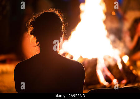 Silhouette der Mann mit dem langen Haar vor dem Feuer sitzen, von hinten während der dunklen Nacht Lagerfeuer gesehen, verschwommenen Hintergrund Stockfoto
