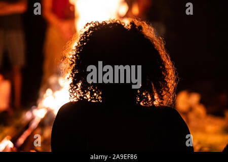 Silhouette einer lockiges Haar Frau in der vor dem Feuer sitzen, von hinten während der dunklen Nacht Lagerfeuer gesehen, verschwommenen Hintergrund Stockfoto
