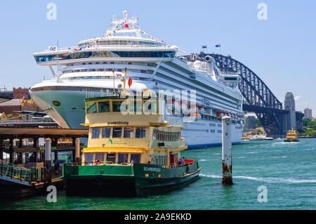 Eine ikonische Fähre im Schatten eines Kreuzfahrtschiffes und die Harbour Bridge - Sydney, NSW, Australien Stockfoto