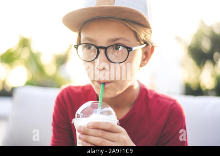 Trendige Junge trinkt ein Smoothie sitzen an der Bar im Freien. Mit Hut Getränke ein kaltes Getränk draußen im Garten jugendlich. Portrait von nachdenklich Teena Stockfoto