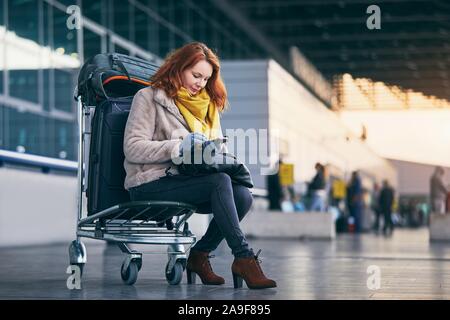 Junge Frau wartet auf Wagen mit Gepäck und mit smart phone gegen Flughafen Terminal Gebäude. Stockfoto